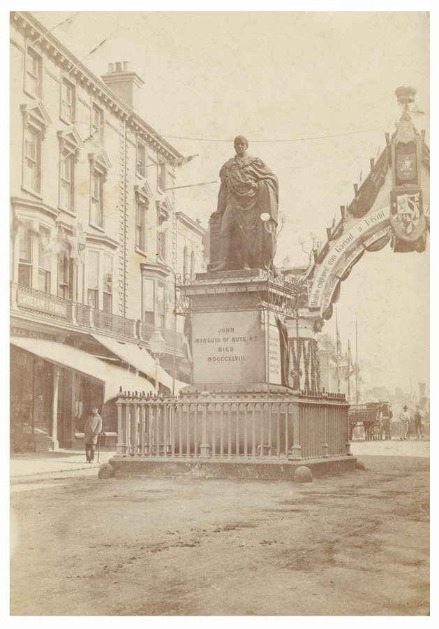 Shows Cardiff High Street decorated for the wedding of the Third Marquess of Bute, John, to Gwendolen Fitzalan-Howard in 1872. Also shown is the statue of the Second Marquess of Bute, John Crichton Stuart (1793-1848), which was unveiled in 1853.